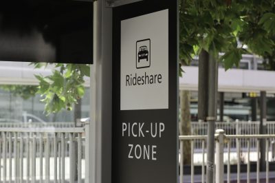 Black and white rideshare pick-up zone sign. Green leaves, metal fence and sign in the background.
